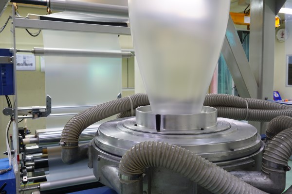 Close-up of a blown film extrusion machine producing plastic film in an industrial setting, featuring rollers, tubing, and a transparent film bubble.